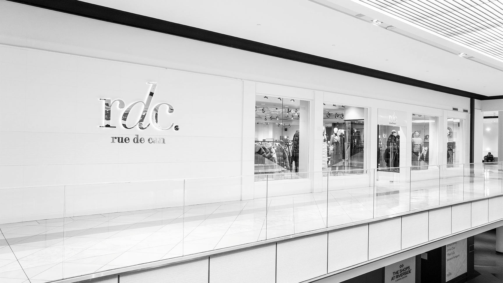 Black and white image of the Rue de Can store located in Hackensack, New Jersey. The storefront features large glass windows displaying clothing items and a modern sign reading 'rdc. rue de can' on the white exterior wall. The store is situated in an upscale shopping mall with a clean, minimalistic design.
